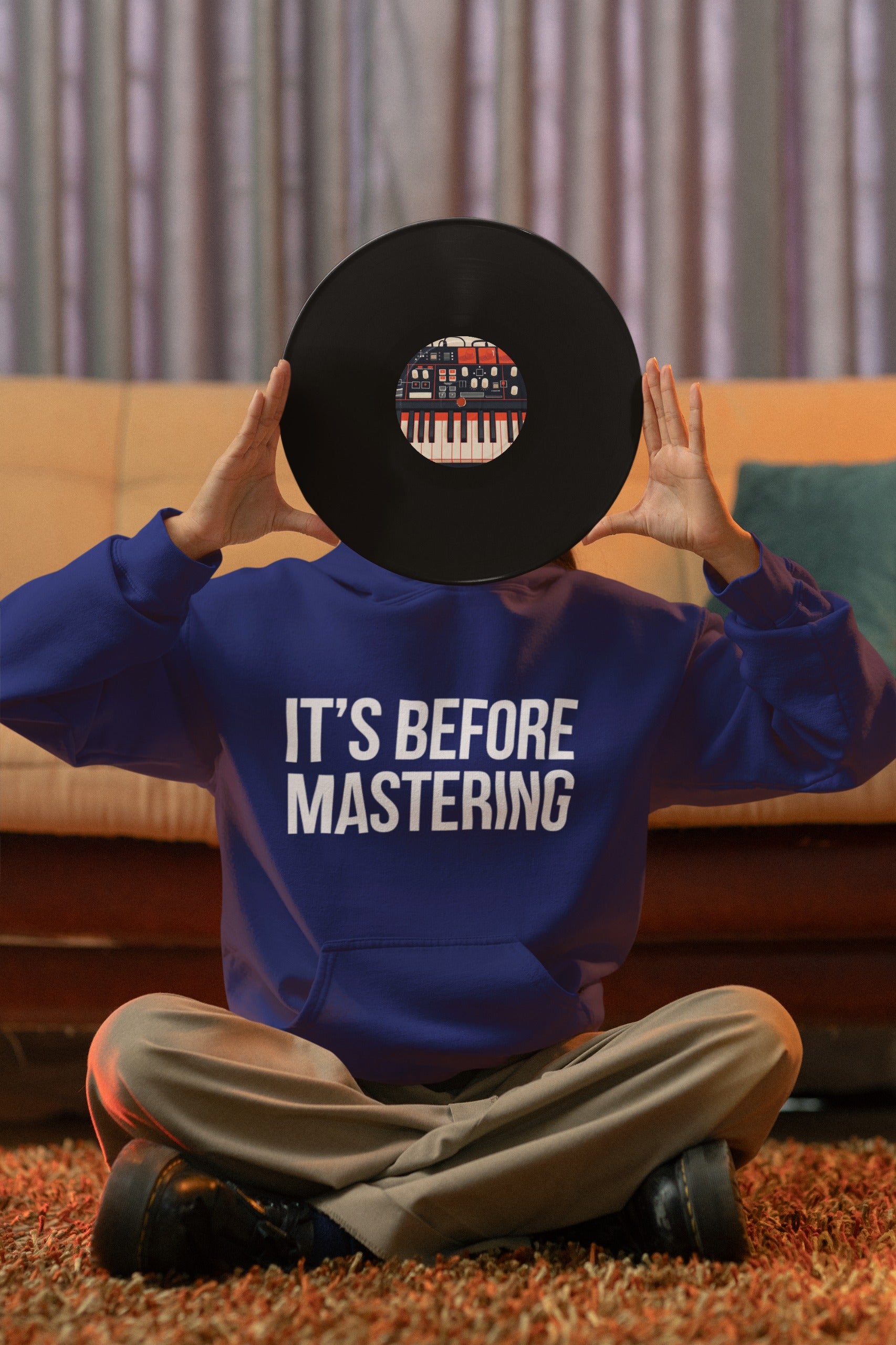Person in a blue sweatshirt that reads "IT'S BEFORE MASTERING," sitting cross-legged on the floor, playfully holding a vinyl record that covers their face against a cozy room background.